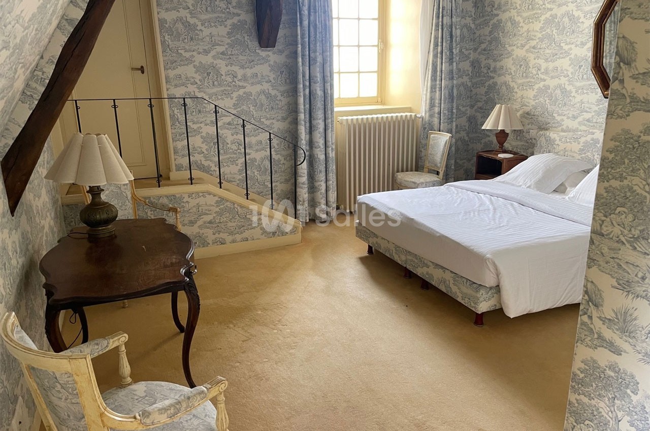 Chambre lumineuse avec lit double, mobilier classique en bois, murs et rideaux à motifs, et fenêtre donnant sur l'extérieur.
