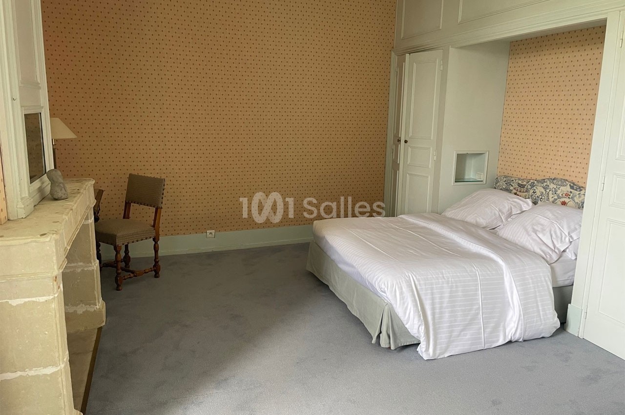 Chambre avec lit double, draps blancs, cheminée en pierre, chaise en bois et murs tapissés de motifs discrets.