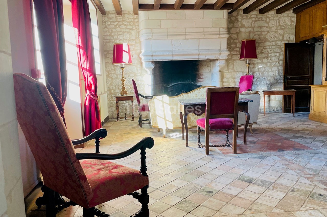 Salle de séjour lumineuse avec cheminée en pierre, meubles anciens et rideaux rouges encadrant des fenêtres.