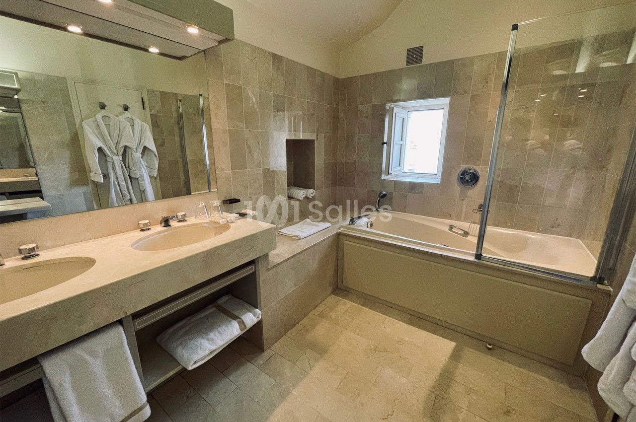 Salle de bain avec baignoire, double vasque, miroir éclairé, peignoirs accrochés et carrelage beige.