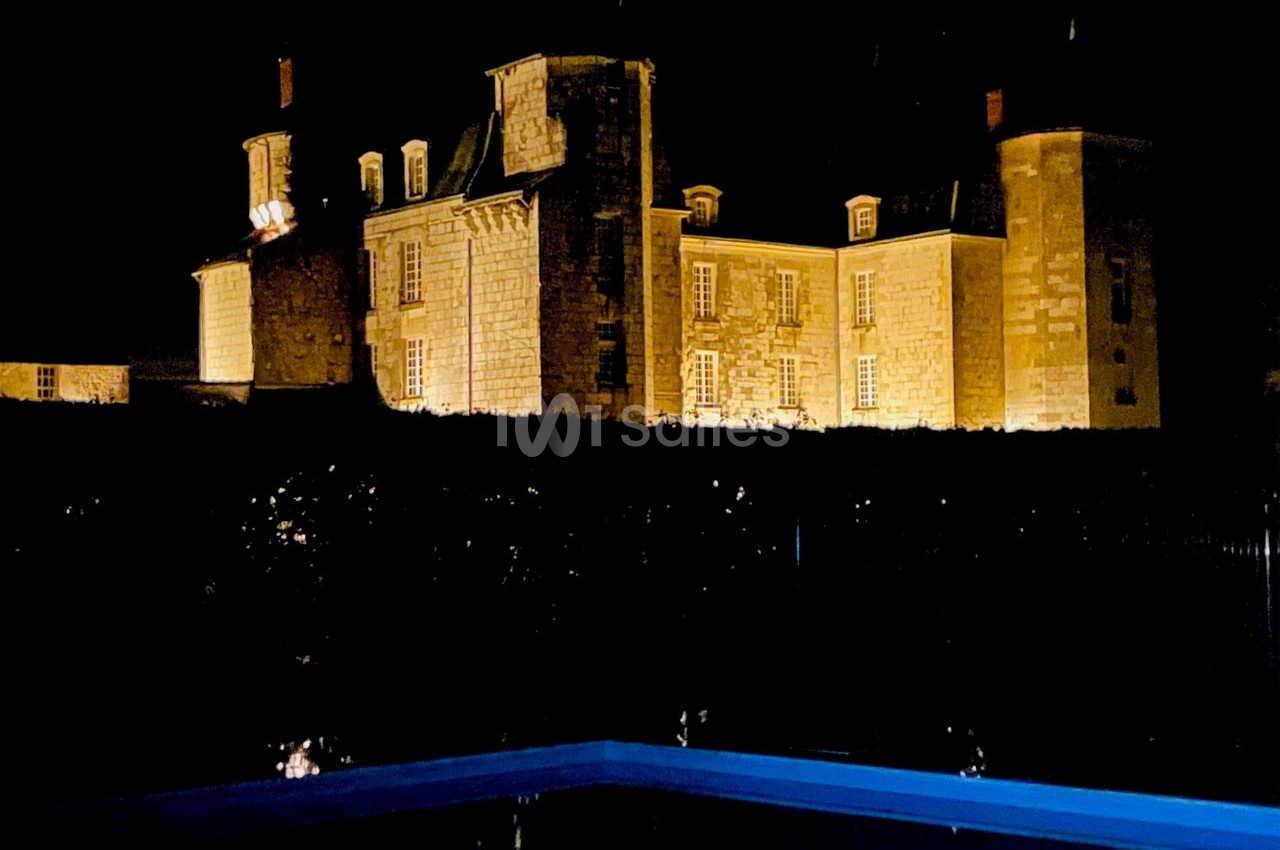 Château en pierre éclairé la nuit, avec une piscine en premier plan reflétant une lueur bleue.