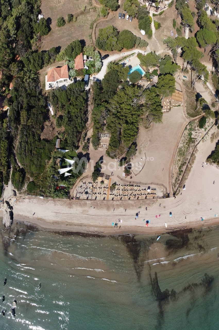Vue aérienne d'une plage bordée de végétation dense, avec des parasols, des bâtiments et une piscine en arrière-plan.