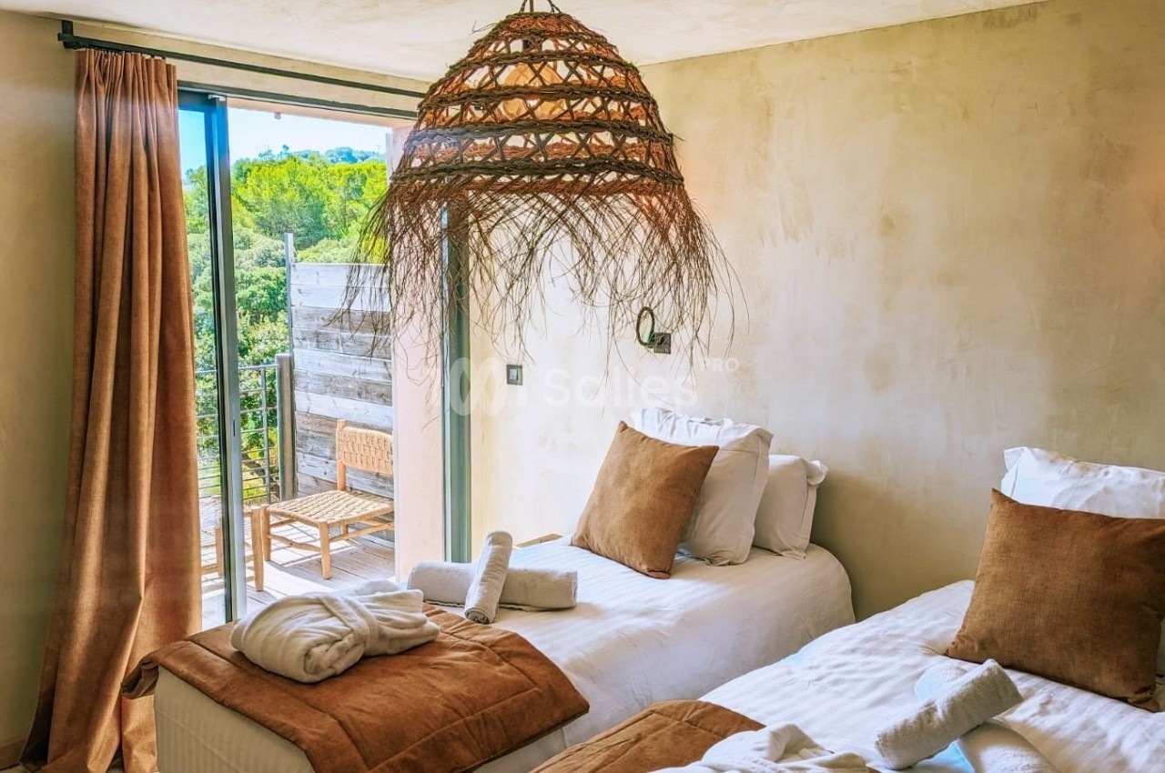 Deux lits simples avec coussins marron dans une chambre lumineuse, ouverte sur une terrasse avec vue sur la végétation.