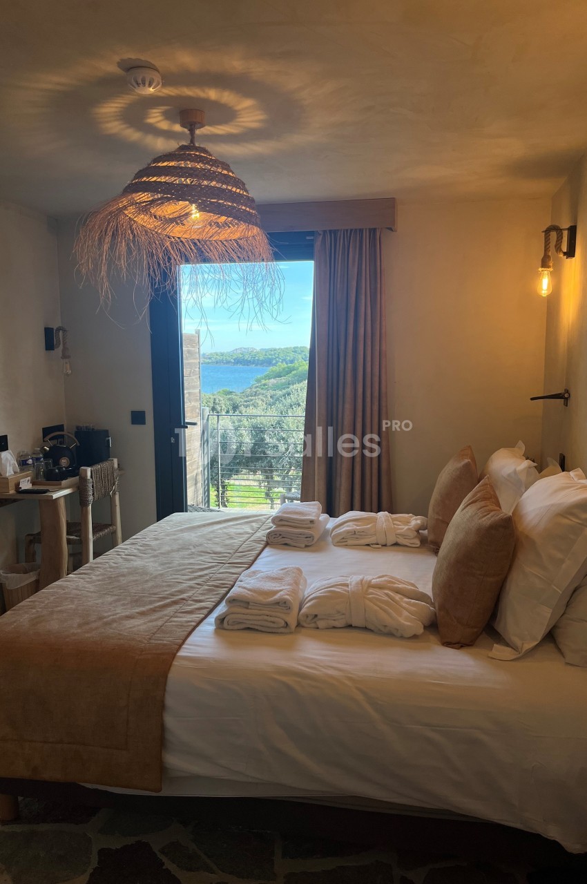 Chambre lumineuse avec lit double, décoration naturelle, vue sur un paysage verdoyant et la mer à travers une porte-fenêtre.