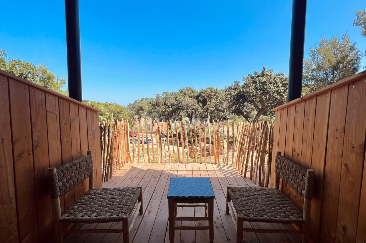 Deux chaises en osier et une table basse sur une terrasse en bois, entourée de palissades et donnant sur un paysage arboré.