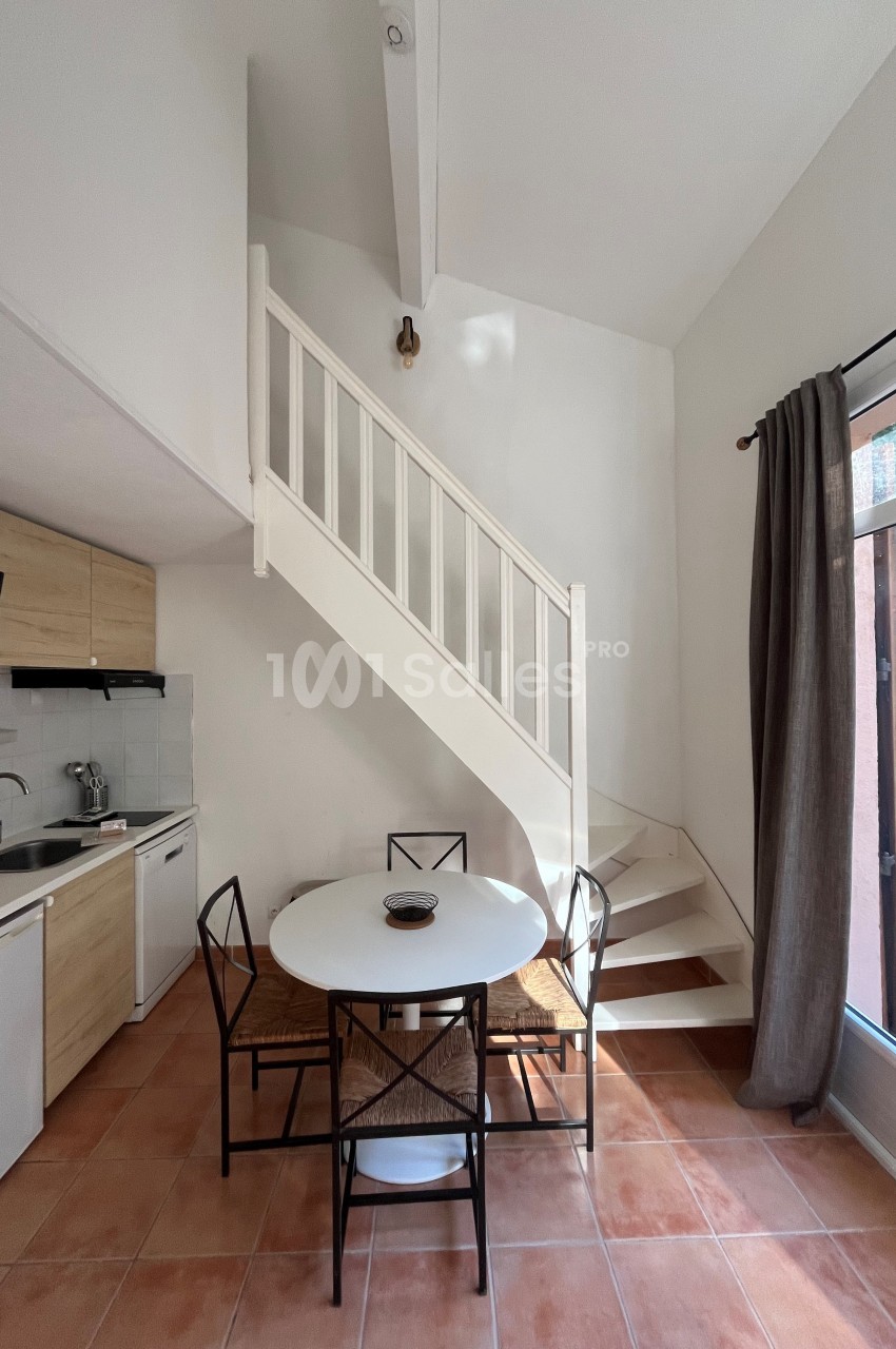 Cuisine ouverte avec table ronde, quatre chaises, escalier blanc, et grande fenêtre avec rideau gris.