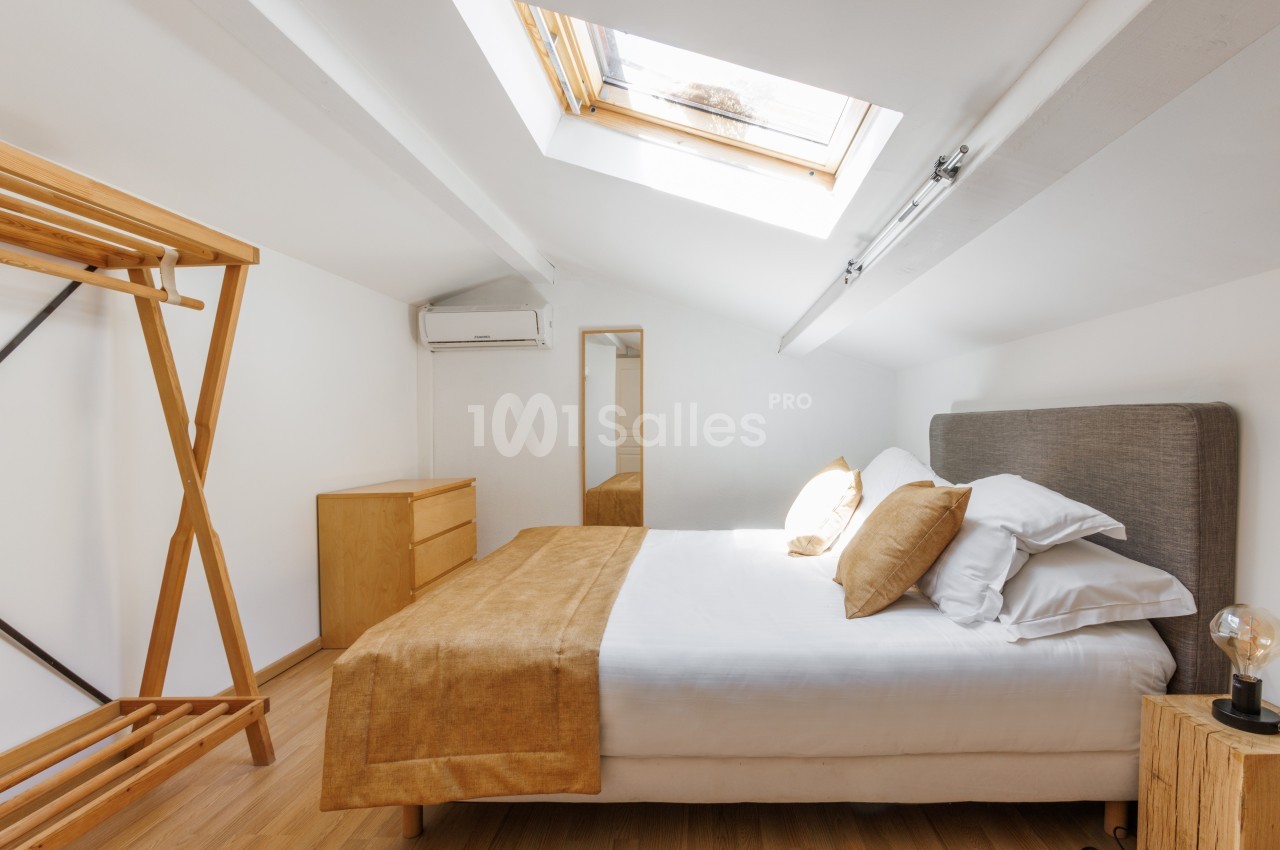 Chambre mansardée lumineuse avec lit double, linge beige, mobilier en bois clair et fenêtre de toit.