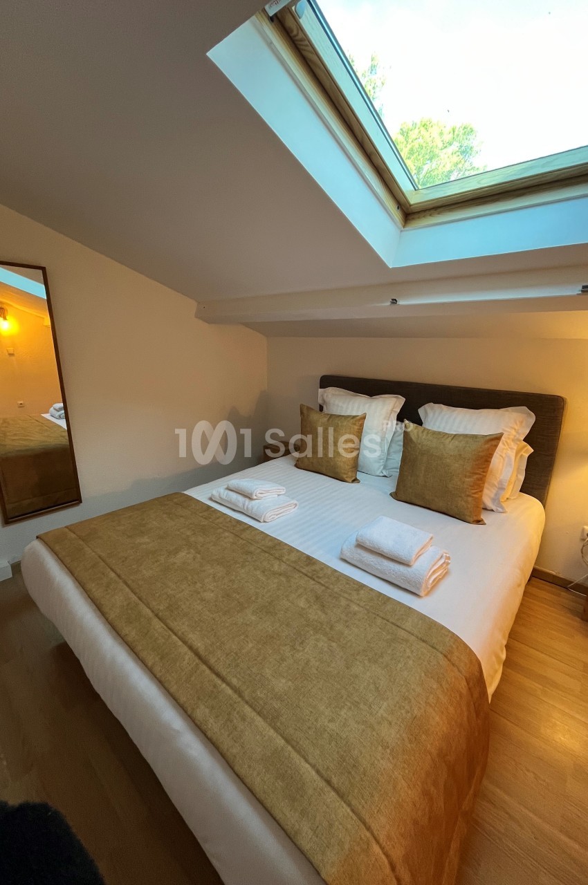 Lit double avec draps blancs et couvre-lit beige, placé sous une fenêtre de toit laissant entrer la lumière naturelle.