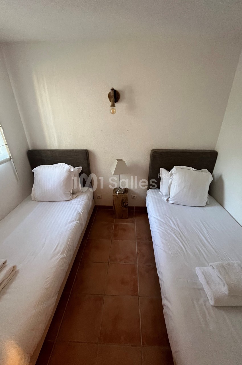 Deux lits simples avec draps blancs, une lampe de chevet centrale et un sol en carrelage dans une chambre lumineuse.