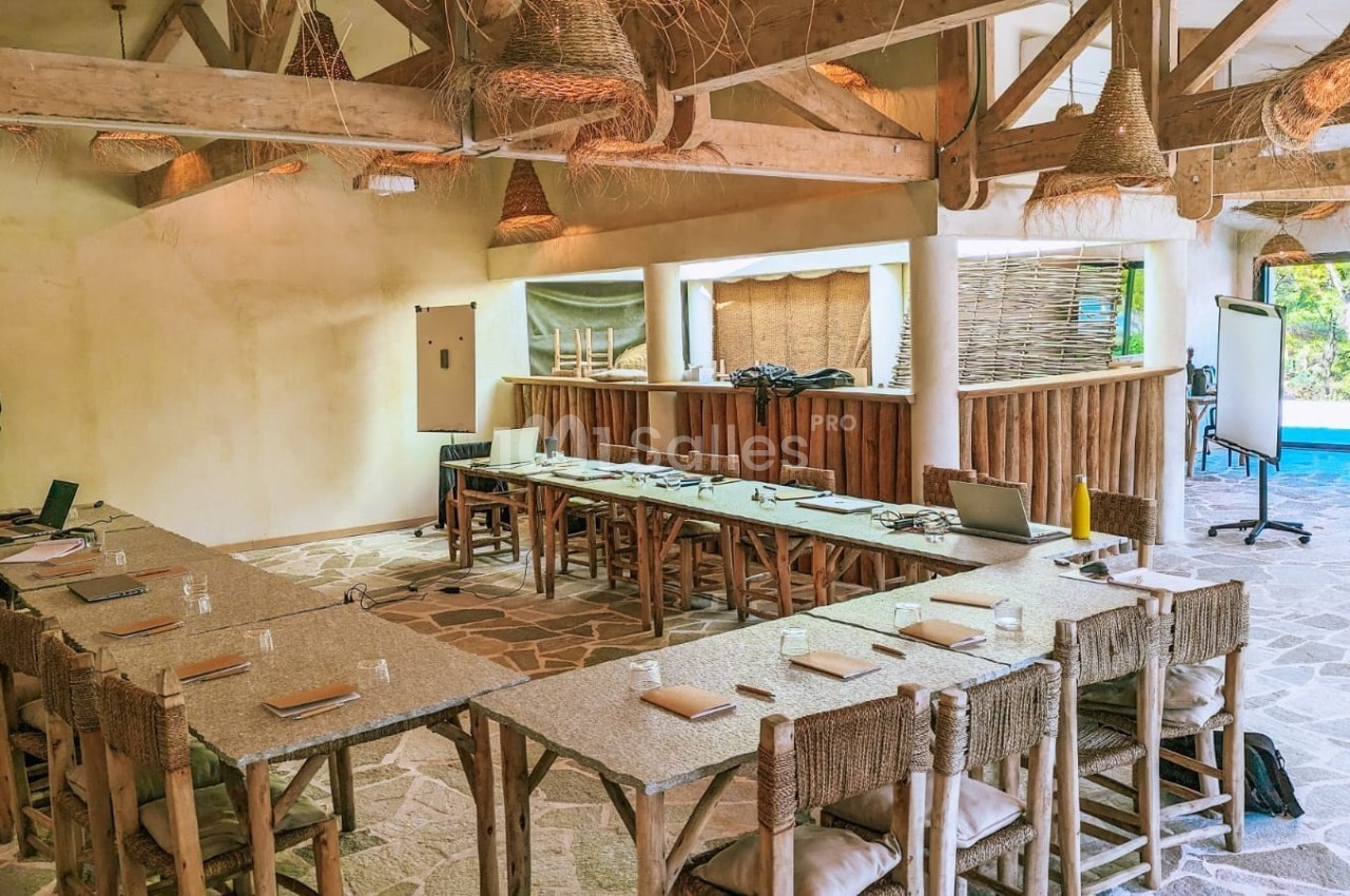 Salle de réunion lumineuse avec tables en U, chaises en bois, décor naturel et vue sur un espace extérieur verdoyant.