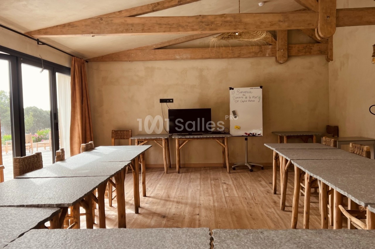 Salle de réunion lumineuse avec tables en U, tableau blanc, écran et poutres apparentes en bois.
