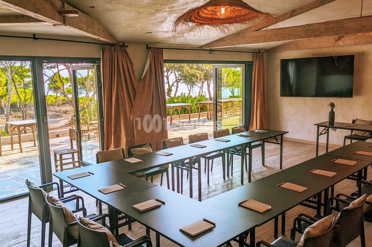 Salle de réunion lumineuse avec grandes baies vitrées, vue sur un espace extérieur arboré et tables disposées en U.