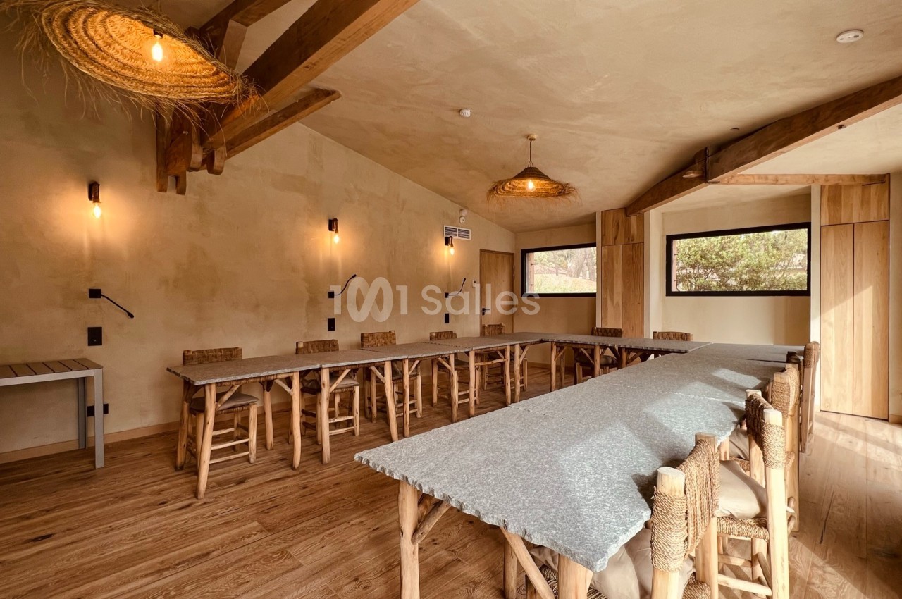 Salle lumineuse avec grandes tables en bois, chaises en osier, poutres apparentes et éclairage suspendu.
