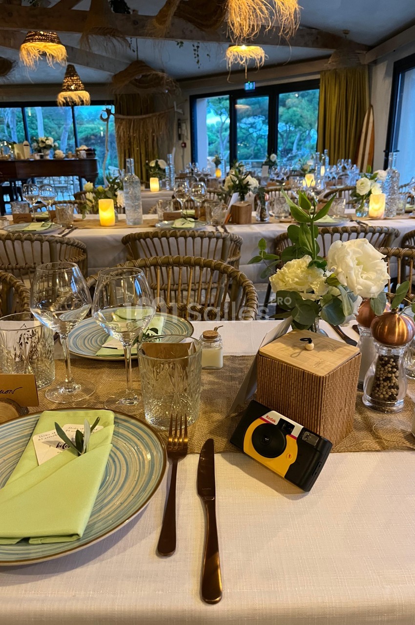 Table dressée pour un repas, avec assiettes, verres, serviettes vertes, décorations florales et appareil photo jetable.