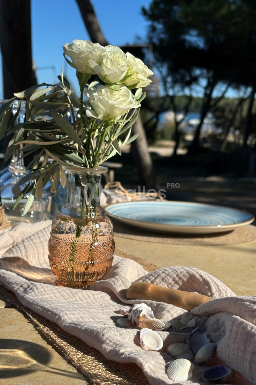 Vase en verre rose avec des fleurs blanches sur une table dressée en extérieur, décorée de coquillages et de tissu beige.