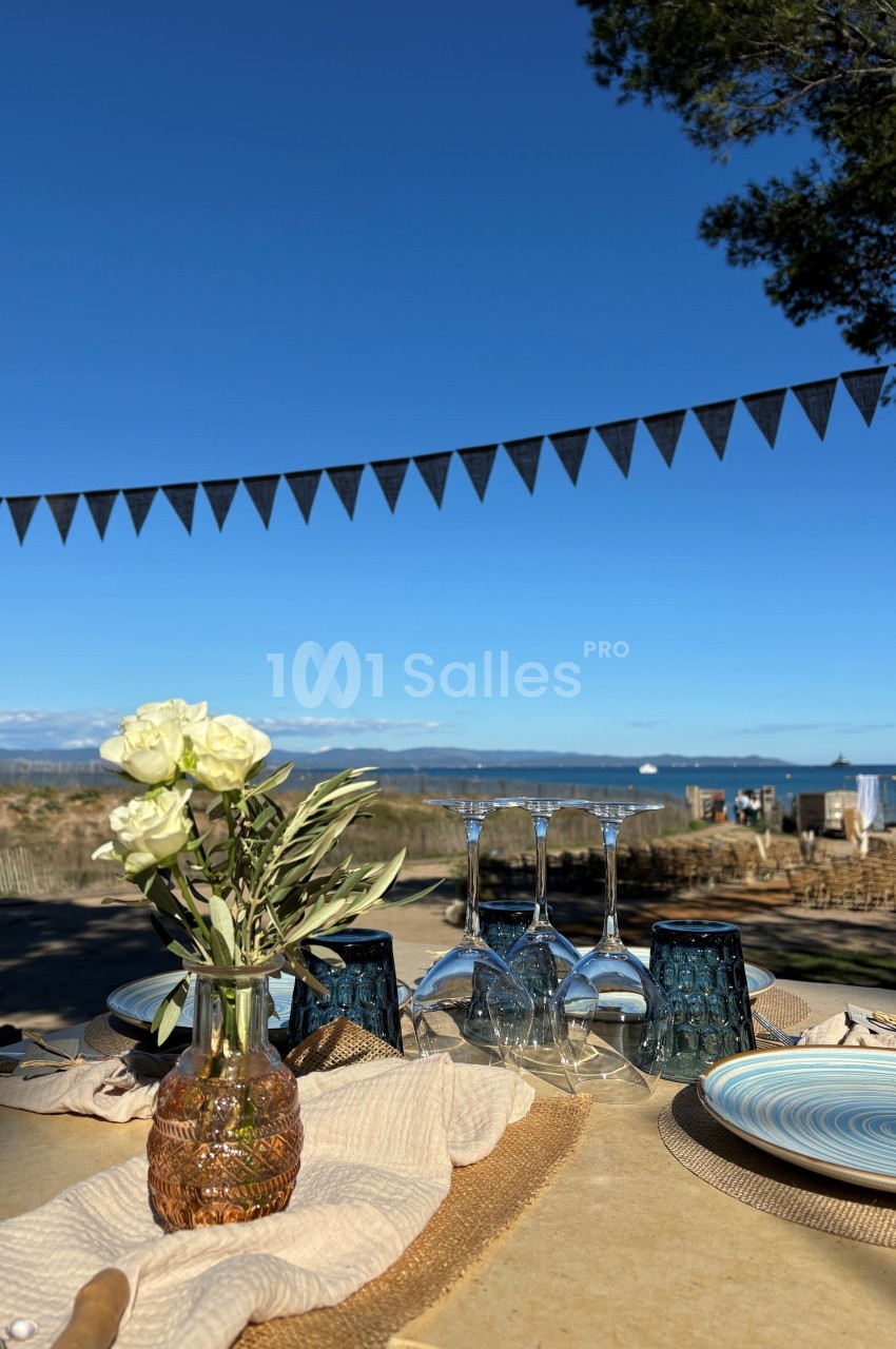 Table extérieure décorée avec des fleurs, vaisselle et verres, avec vue sur la mer et un ciel bleu dégagé.