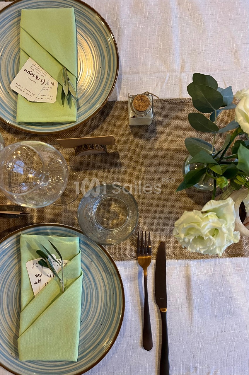 Table dressée avec assiettes en céramique, serviettes vertes pliées, couverts dorés, verres et décoration florale blanche.