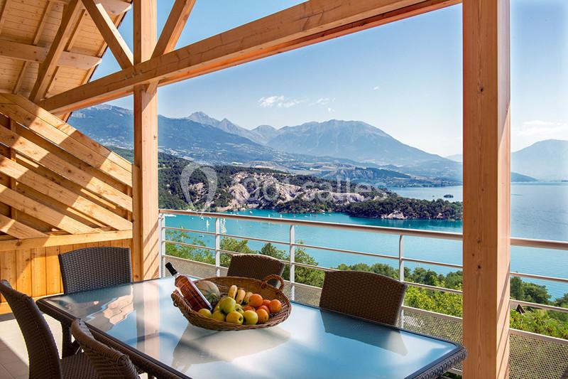 Vue sur un lac entouré de montagnes depuis une terrasse en bois avec une table en verre et un panier de fruits.