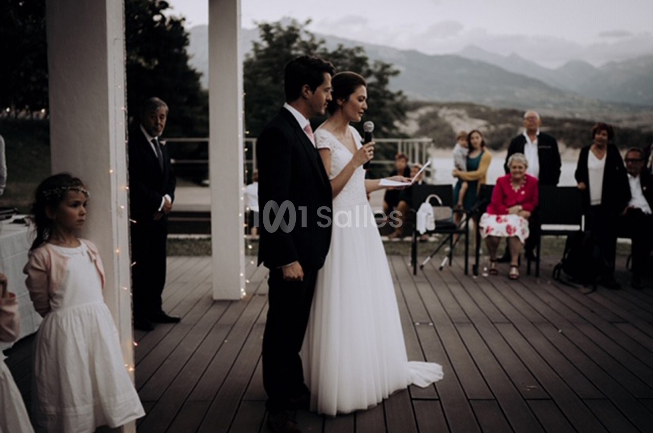 Un couple en tenue de mariage prononce un discours sur une terrasse en présence d'invités assis et debout.
