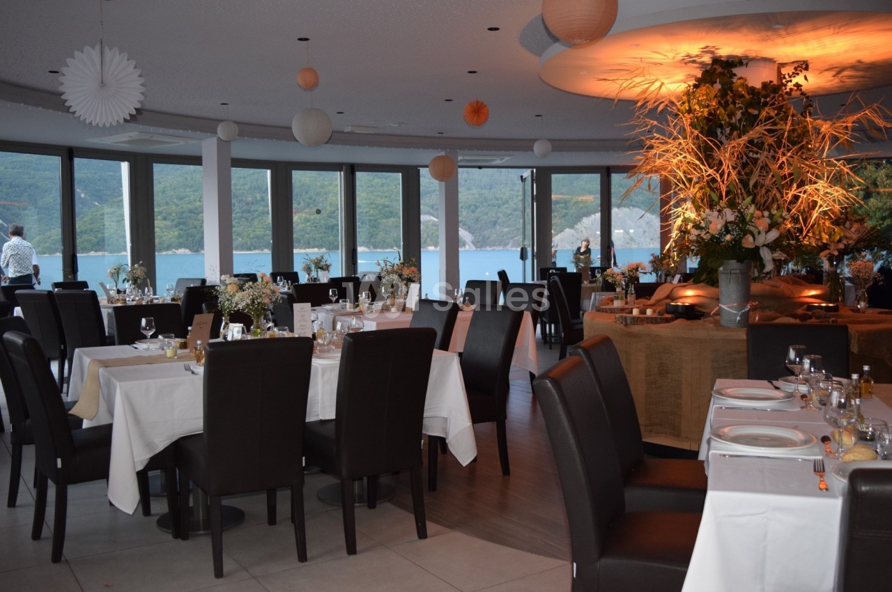 Salle de restaurant élégante avec grandes baies vitrées donnant sur un paysage de lac et montagnes, tables dressées.