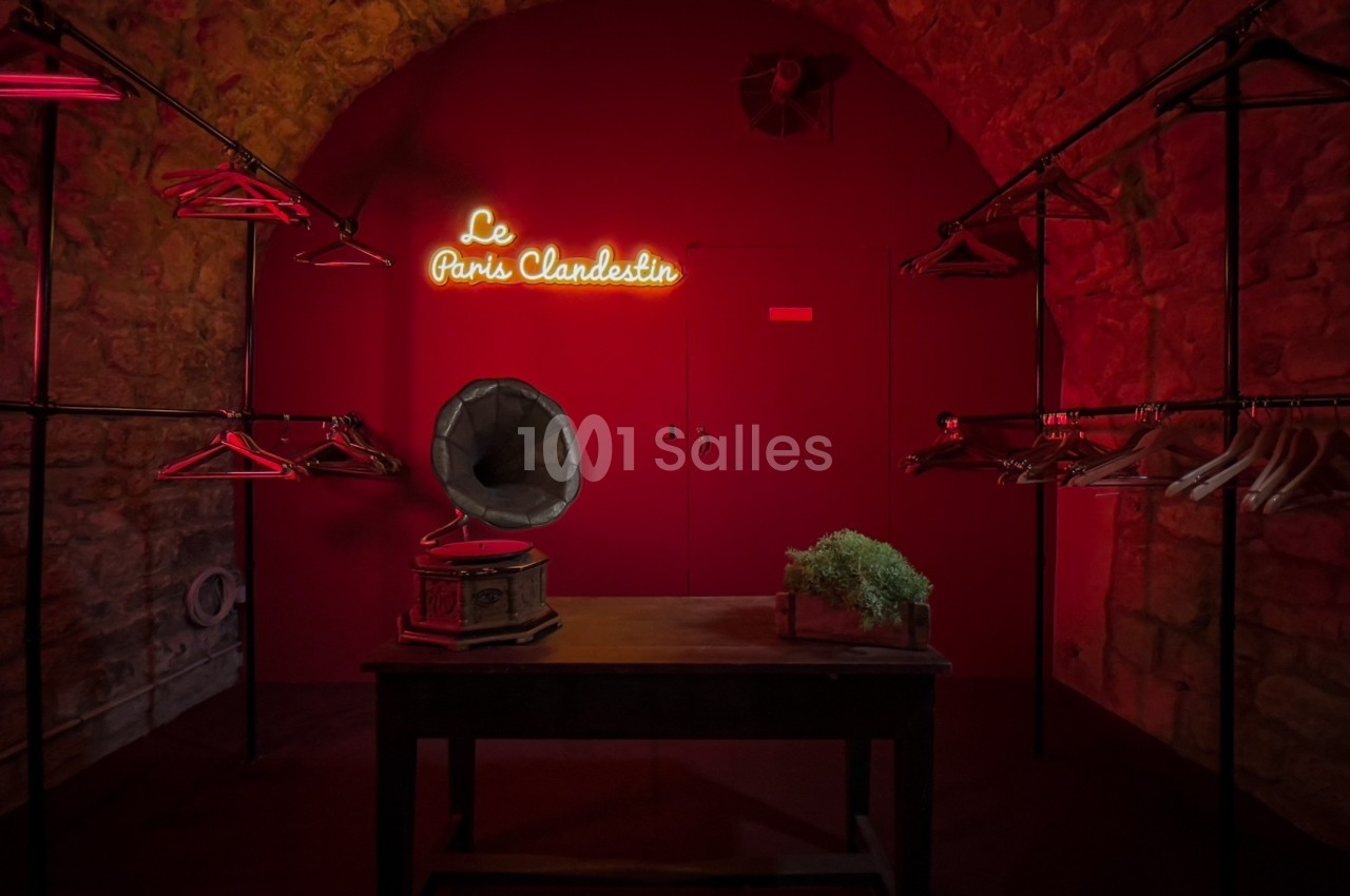 Salle en pierre éclairée de rouge avec un gramophone sur une table, des portants vides et une enseigne lumineuse.
