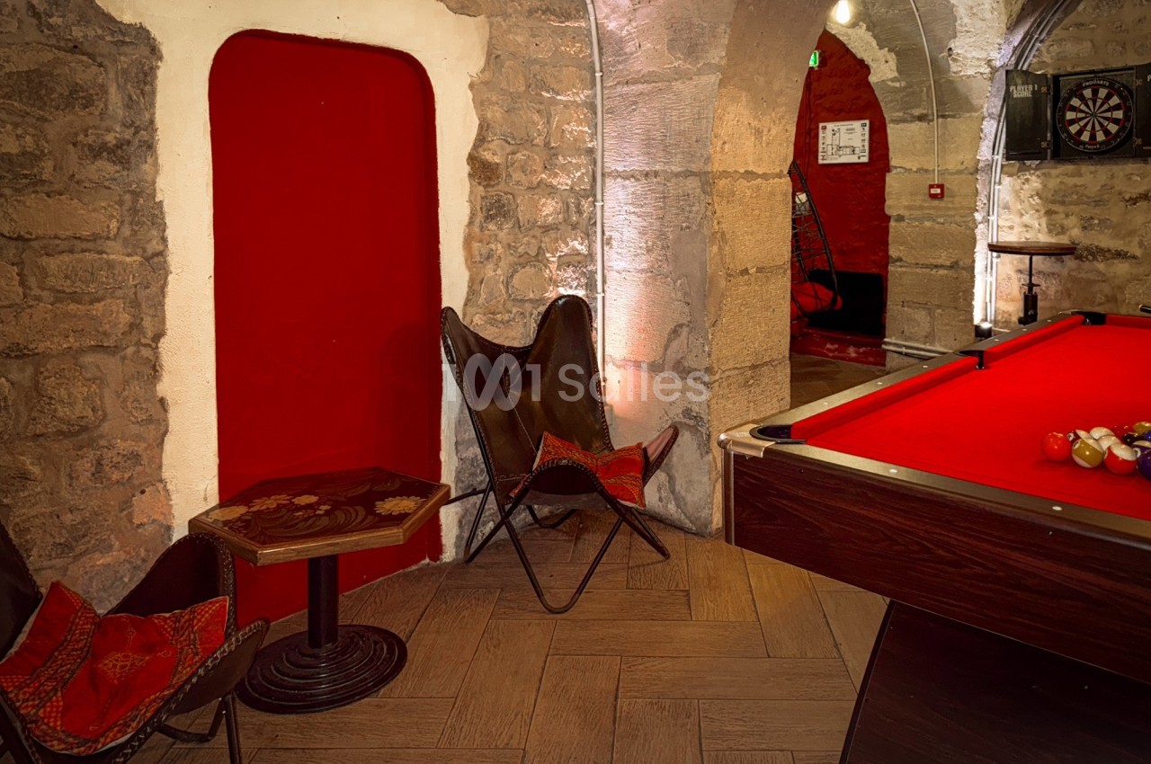 Salle voûtée en pierre avec un billard rouge, deux fauteuils en cuir et une cible de fléchettes au fond.