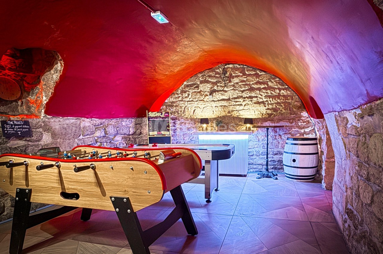 Salle voûtée en pierre avec baby-foot, tonneau en bois, éclairage chaleureux et murs peints en rouge.