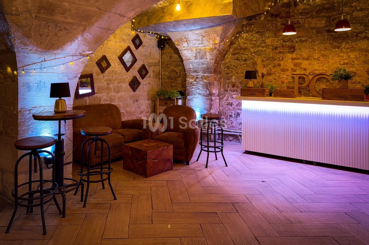 Salle voûtée en pierre avec un bar éclairé, des fauteuils en velours, des tabourets et une ambiance lumineuse chaleureuse.
