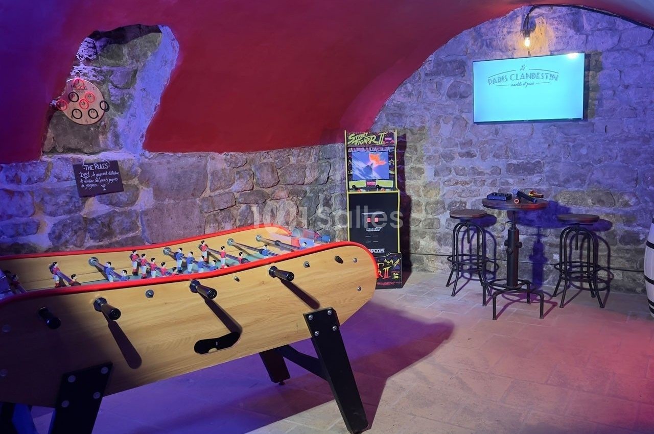 Salle voûtée en pierre avec baby-foot, borne d'arcade, table haute et écran mural allumé.
