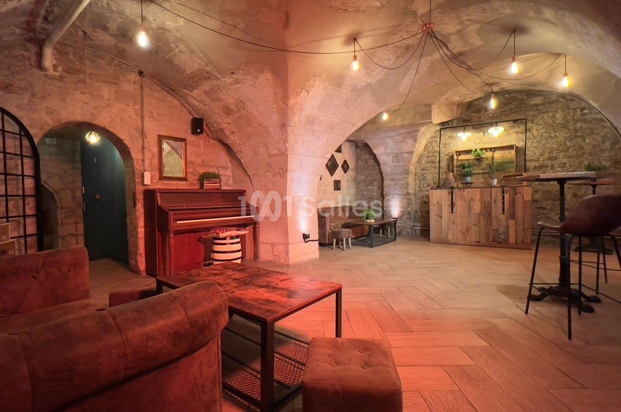 Salle voûtée en pierre avec éclairage chaleureux, mobilier en bois et cuir, piano et comptoir en arrière-plan.