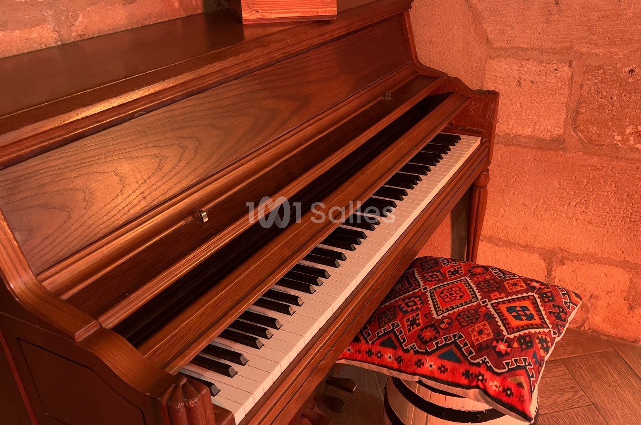 Piano droit en bois avec un tabouret recouvert d'un coussin à motifs colorés, posé dans une pièce aux murs en pierre.