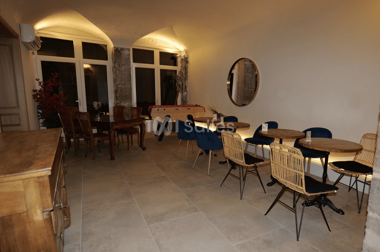 Salle lumineuse avec tables, chaises variées, baby-foot, miroir rond et murs en pierre apparente.