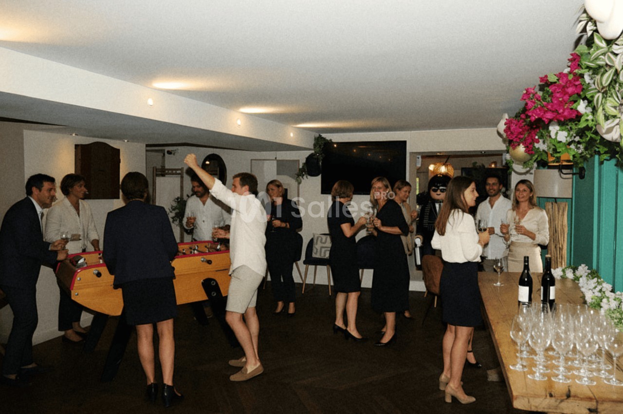Des personnes discutent et jouent au baby-foot dans une salle lumineuse décorée de fleurs et de tables dressées.