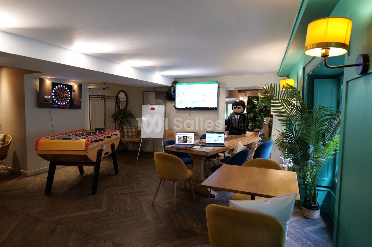 Salle de détente avec baby-foot, fléchettes, tables, chaises, plantes et écrans allumés.