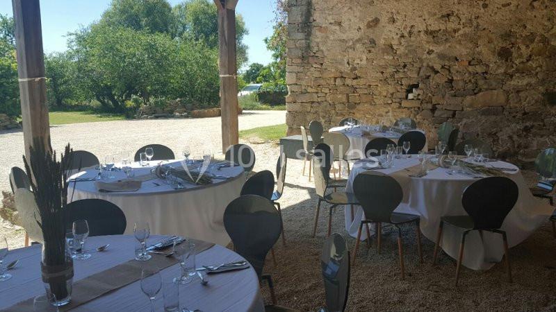 Tables rondes dressées avec nappes blanches et chaises modernes dans un espace extérieur semi-ouvert près d'un mur en pierre.