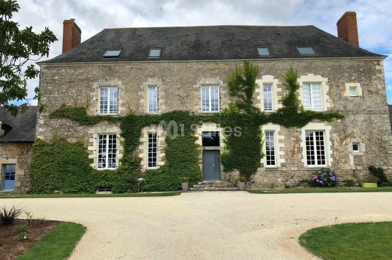 Façade d'une grande maison en pierre avec des murs partiellement recouverts de lierre, entourée d'une allée et de pelouses.