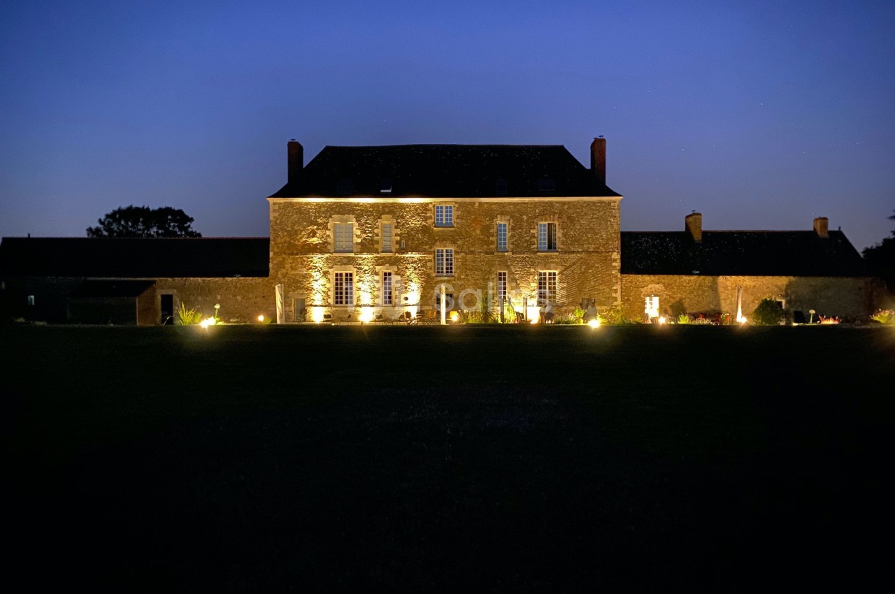 Façade d'une grande maison en pierre éclairée la nuit, entourée d'un jardin sombre.