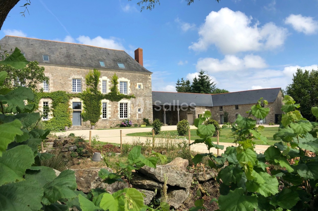 Bâtisse en pierre avec volets blancs, entourée de verdure et d'un jardin, sous un ciel bleu parsemé de nuages.