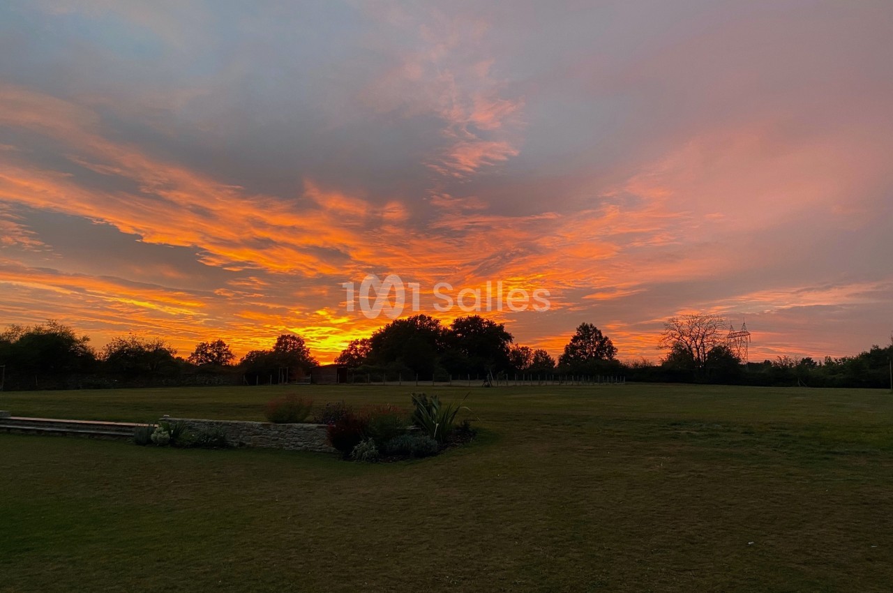 Ciel aux teintes orange et rose au coucher du soleil, avec un grand espace vert et des arbres à l'horizon.