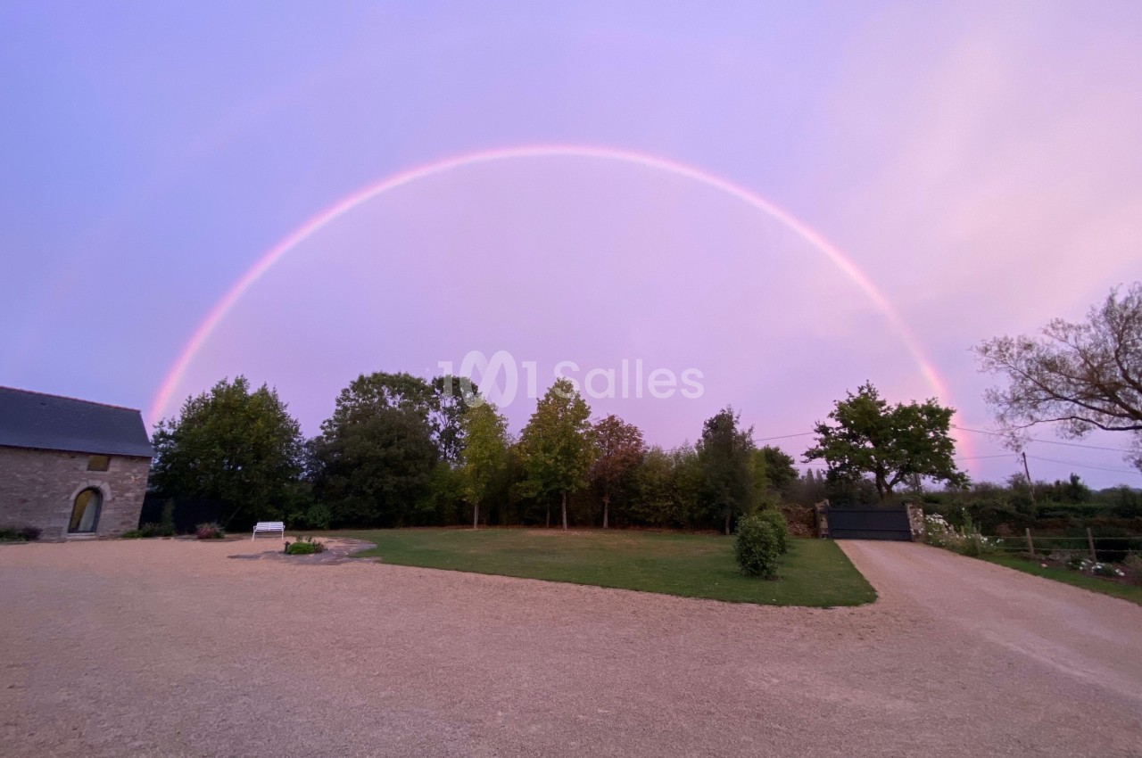 Un arc-en-ciel complet dans un ciel rose au-dessus d'une cour avec pelouse, arbres et bâtiments en arrière-plan.