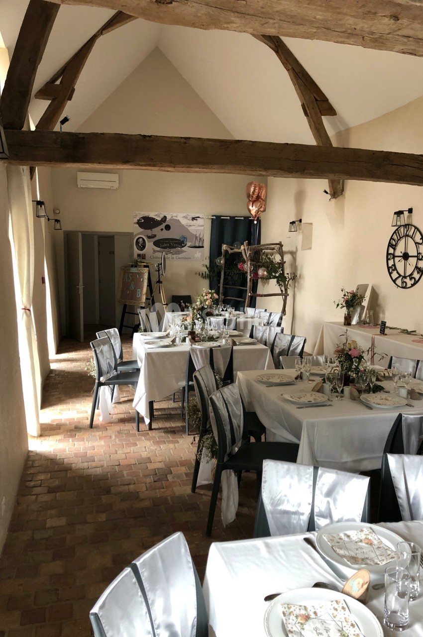 Salle de réception avec tables dressées, décoration florale et poutres apparentes sous un plafond en pente.