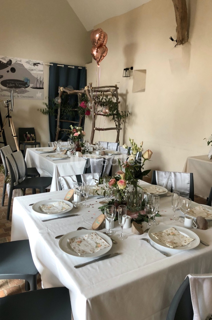 Salle décorée pour un repas festif, avec tables dressées, nappes blanches, fleurs et ballons roses en arrière-plan.