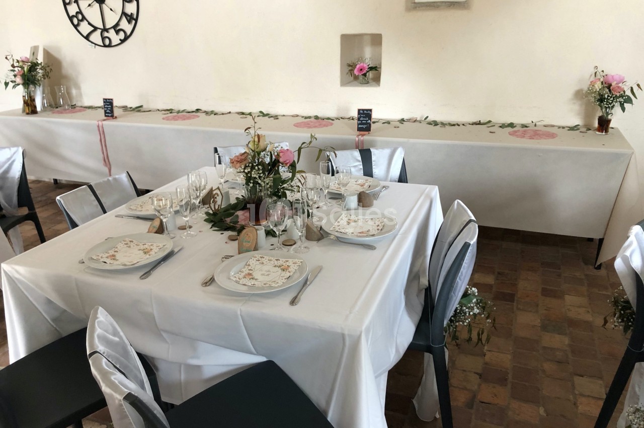 Table dressée pour un repas avec nappes blanches, vaisselle élégante, décor floral et chaises décorées.