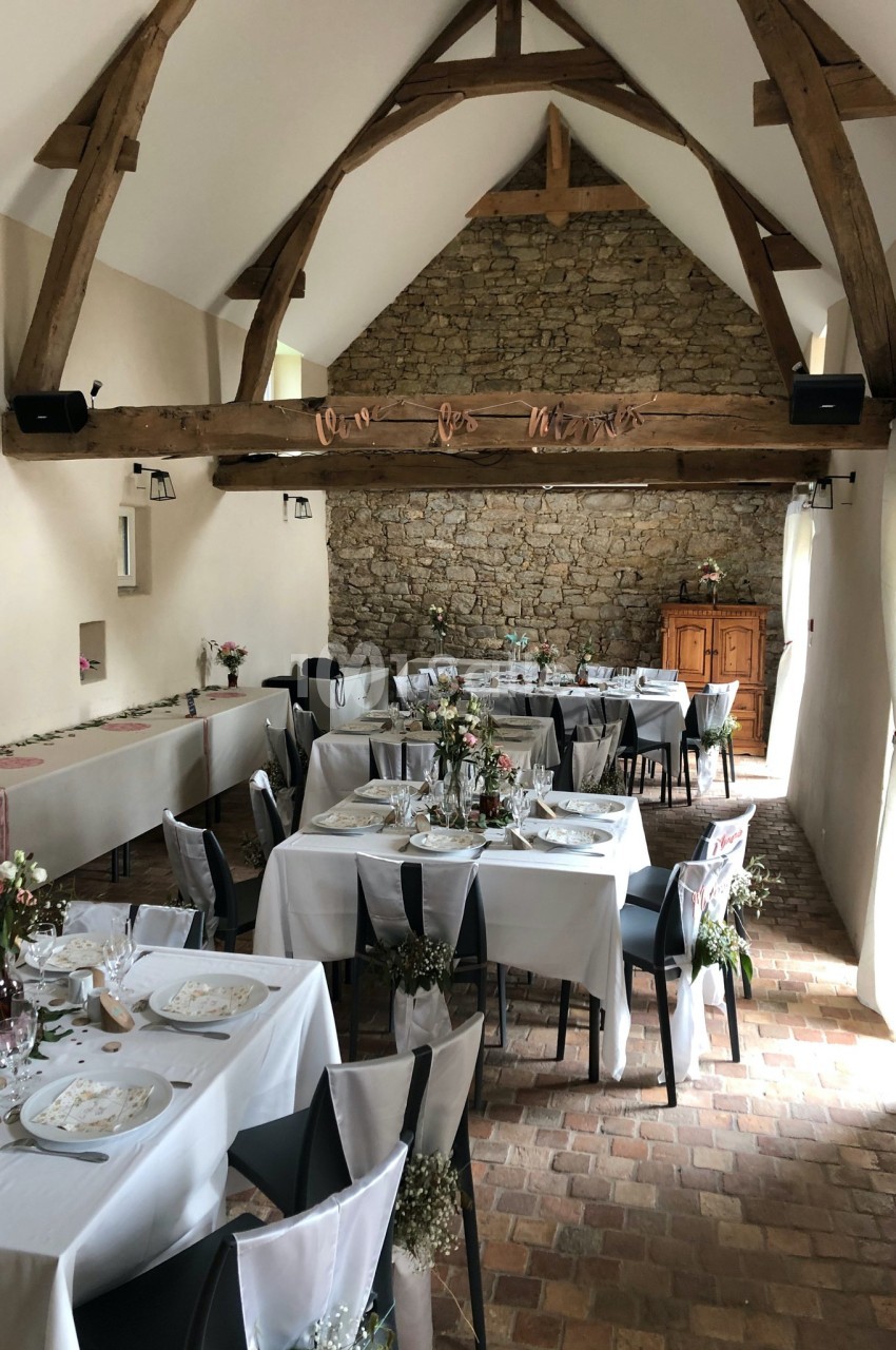 Salle de réception décorée pour un événement, avec tables dressées, poutres apparentes et mur en pierre.