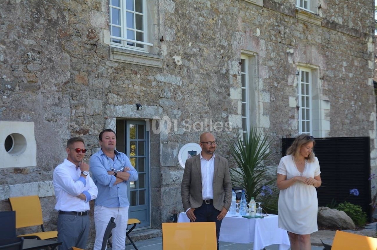 Quatre personnes debout devant un bâtiment en pierre, avec des chaises et une table dressée à proximité.