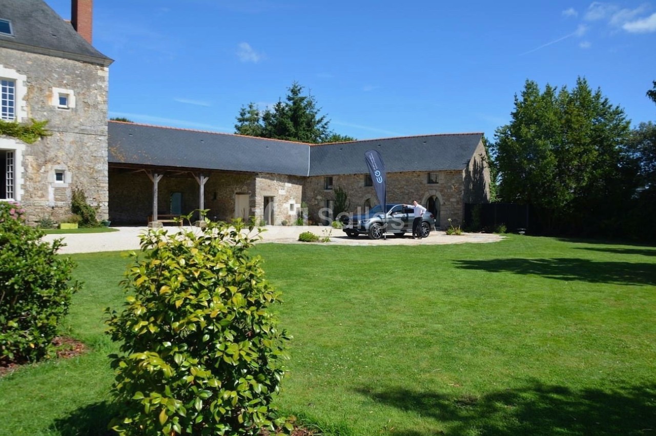 Cour d'une maison en pierre avec pelouse, voiture garée près d'une bannière et arbres en arrière-plan.