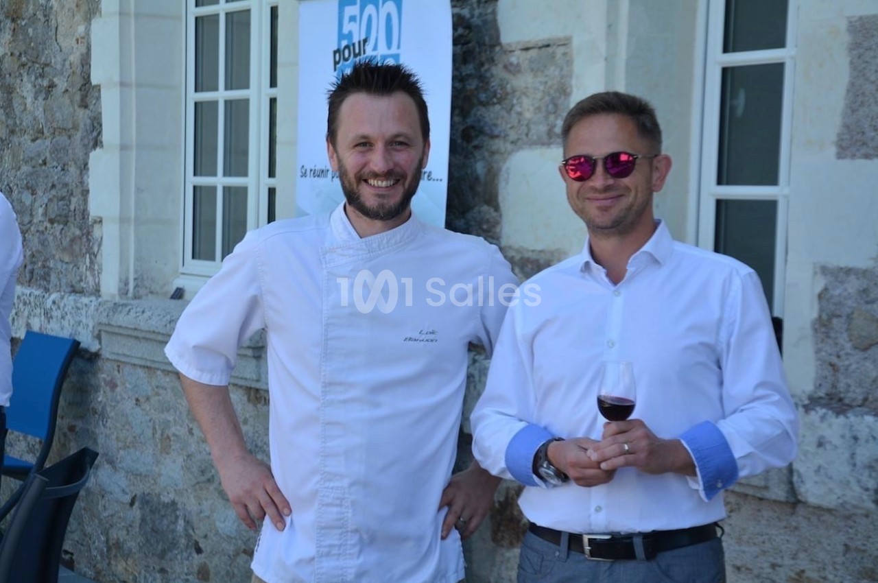 Deux hommes souriants, l'un en tenue de chef, l'autre en chemise blanche, tenant un verre de vin, devant un bâtiment en…