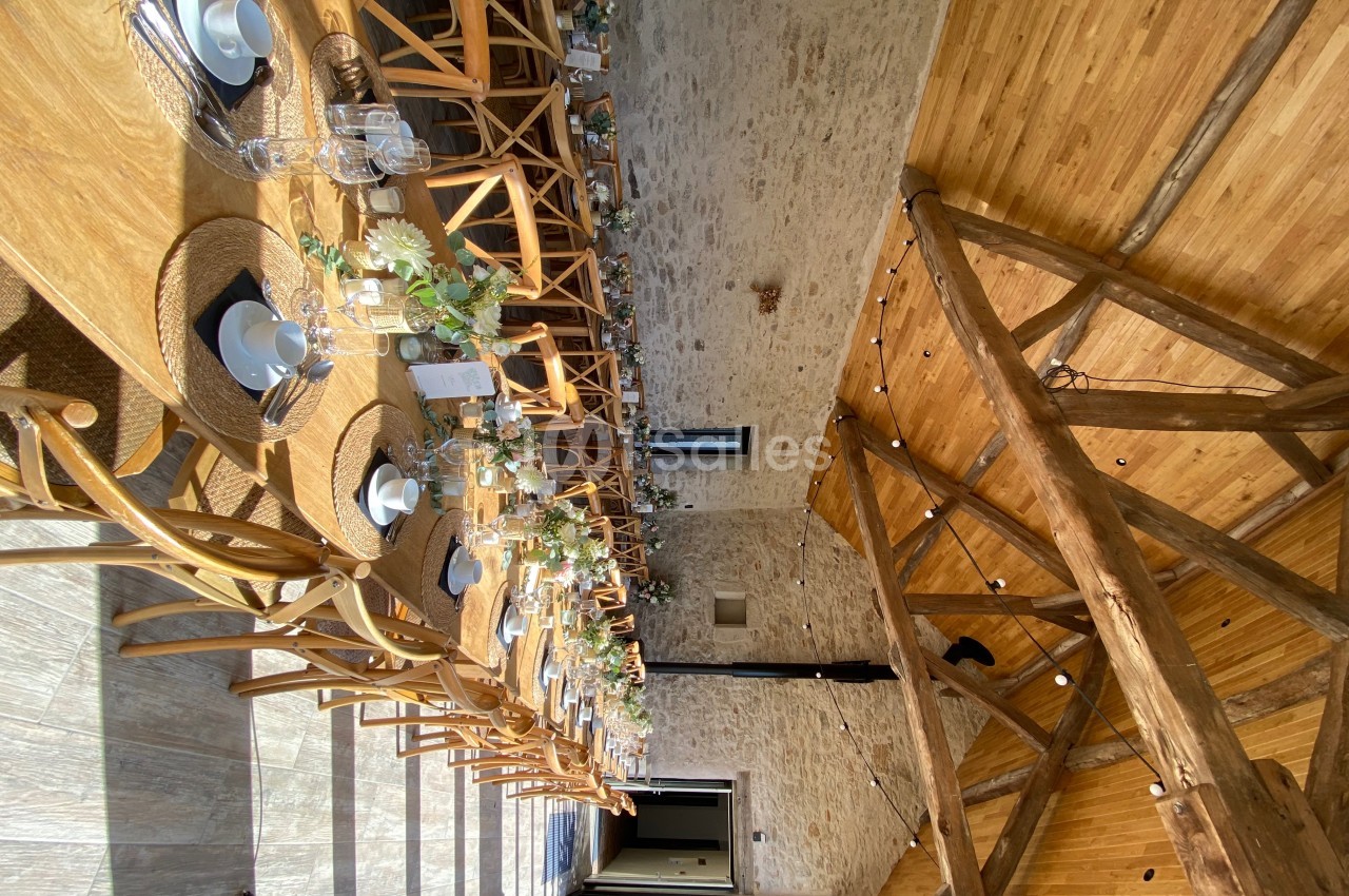 Salle en pierre avec de grandes tables en bois décorées de fleurs et de vaisselle, éclairée par une lumière naturelle.