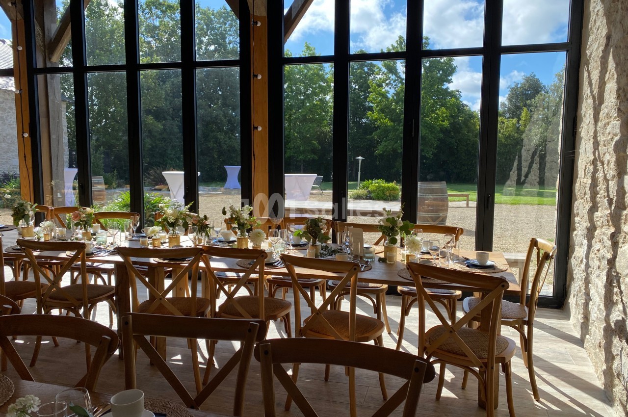 Salle lumineuse avec de grandes baies vitrées, tables décorées pour un repas, vue sur un jardin arboré.