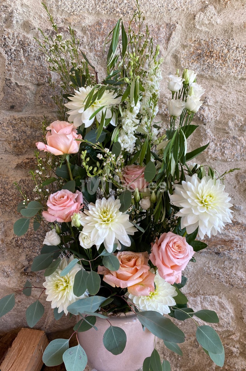 Arrangement floral avec roses roses, dahlias blancs et feuillage vert dans un vase rose, posé devant un mur en pierre.